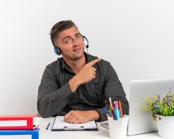 young confident blonde office worker man on headphones sits at desk with office tools using laptop points at side isolated on white background with copy space